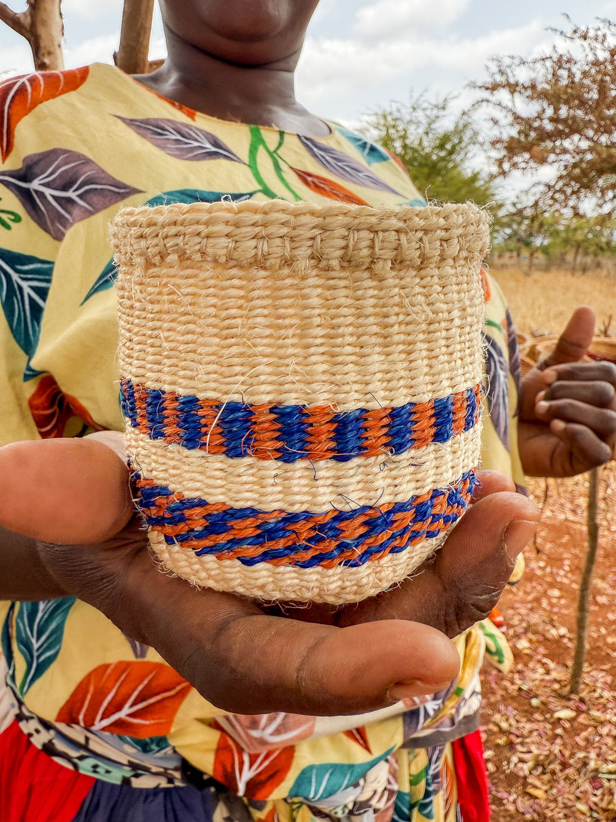 A person in a colorful, leaf-patterned shirt holds out a Thick Weave Sisal Basket with blue and orange stripes. In the background, dry grass and trees are visible beneath a partly cloudy sky.