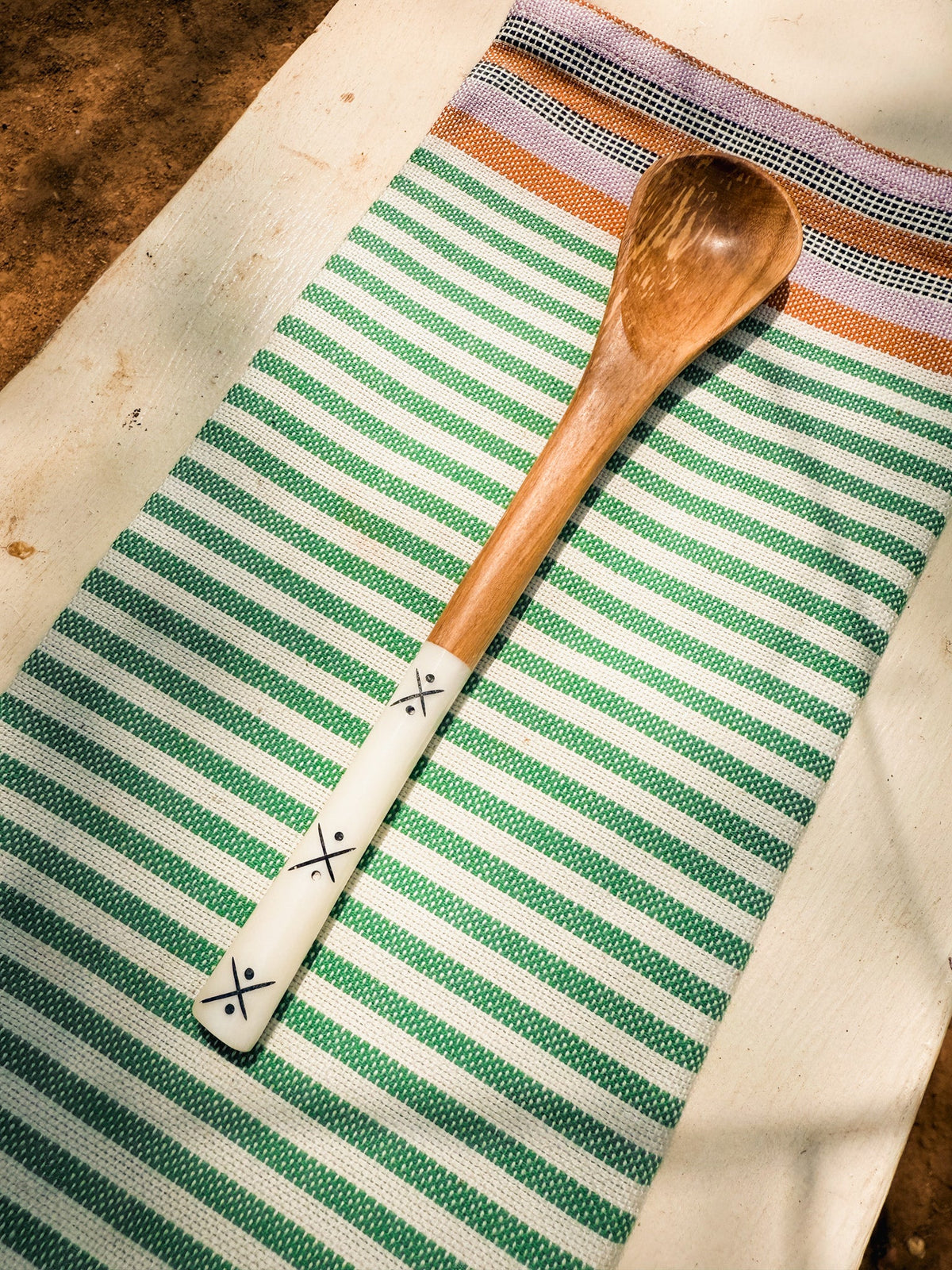 The Olive Wood Sugar & Honey Spoon, handmade with a white handle featuring black X patterns, is displayed on a green and white striped towel—a stunning Kenyan artisan utensil.