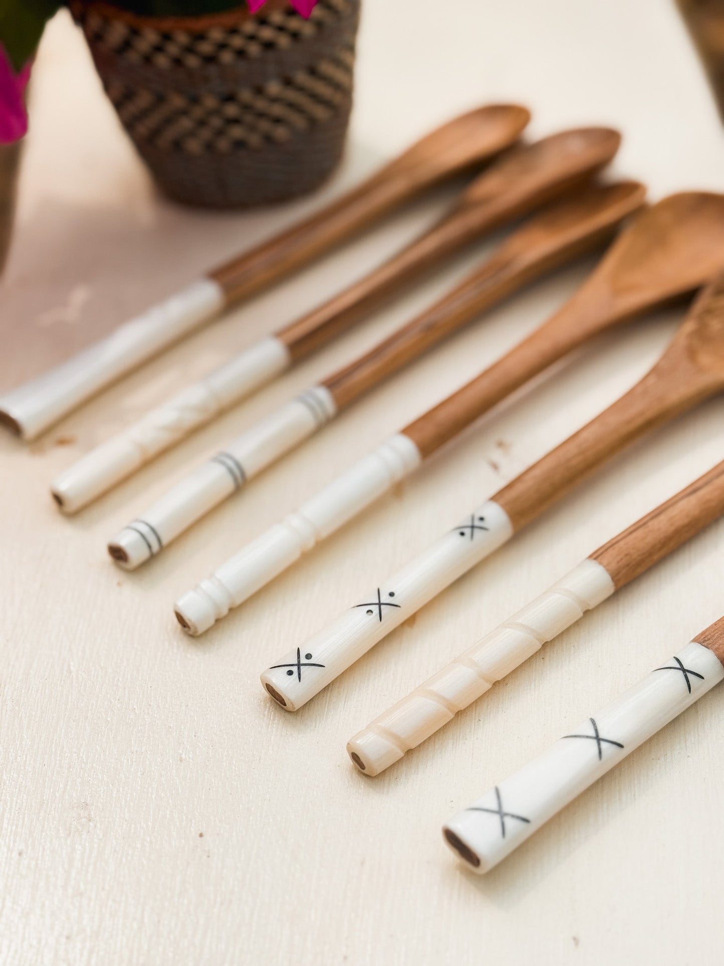 A set of six Olive Wood Sugar & Honey Spoons with white handles—some decorated with black X patterns—are arranged on a light surface. A woven basket holding a plant appears in the background, highlighting these versatile Kenyan artisan utensils.