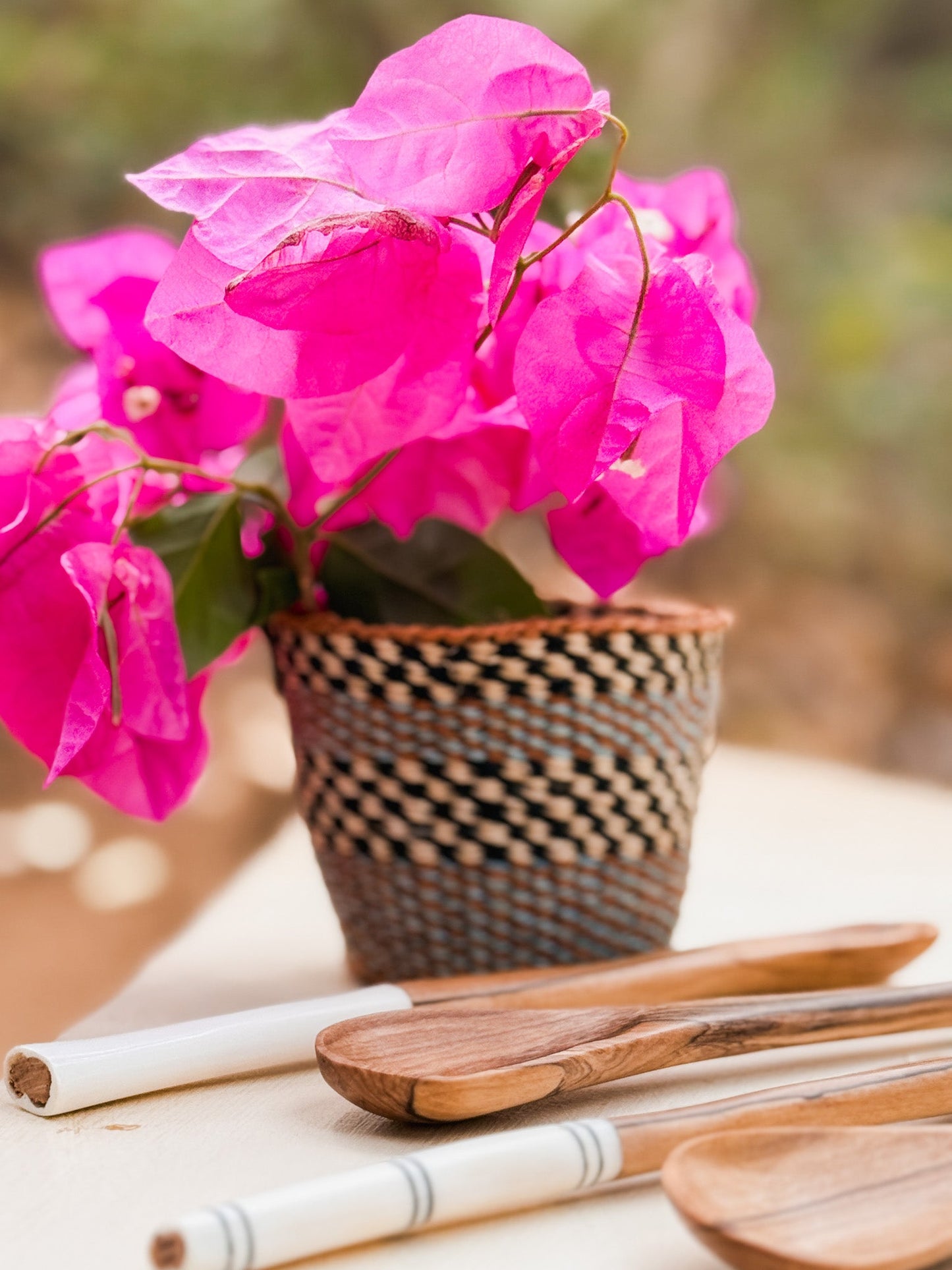 A woven basket with bright pink bougainvillea sits on a table next to Olive Wood Sugar & Honey Spoons and Kenyan artisan utensils. The softly blurred background highlights the natural beauty of these handmade pieces.
