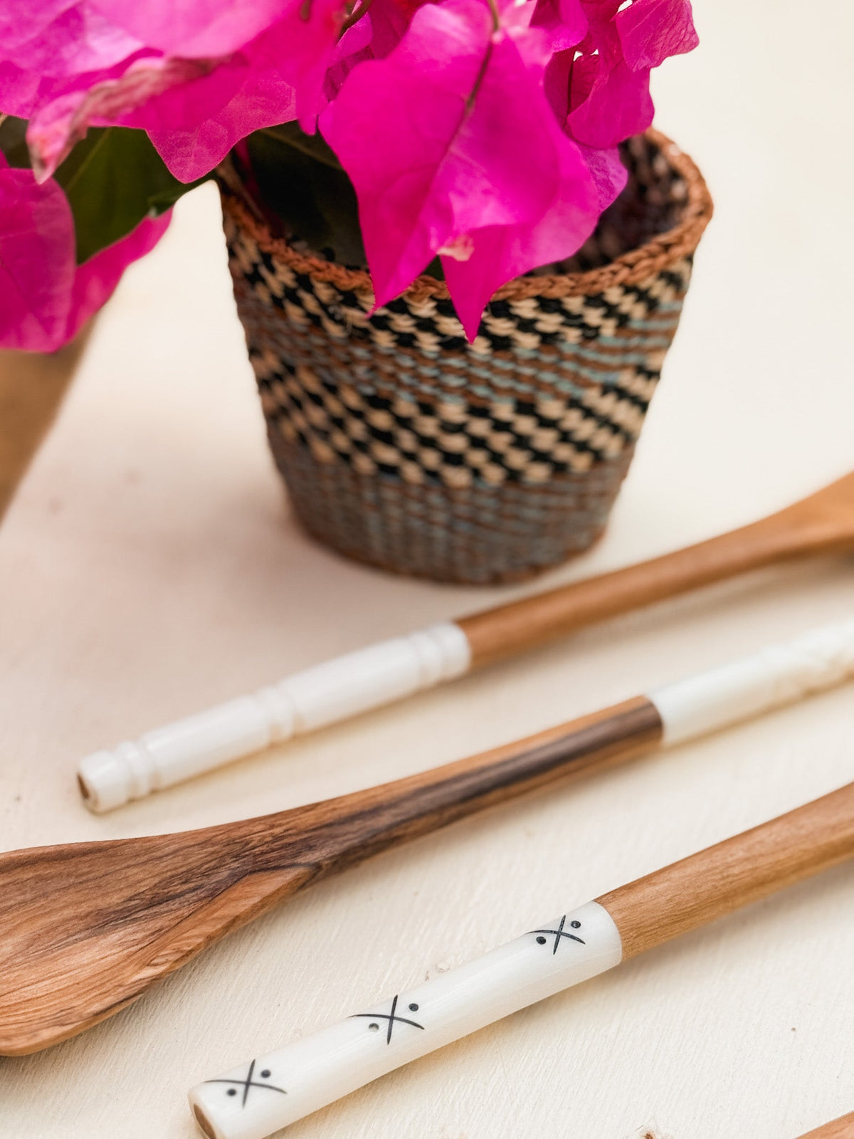 Two Kenyan artisan Olive Wood Sugar & Honey Spoons, featuring white handles with simple decorative patterns, rest on a light surface near a woven basket of bright pink flowers.