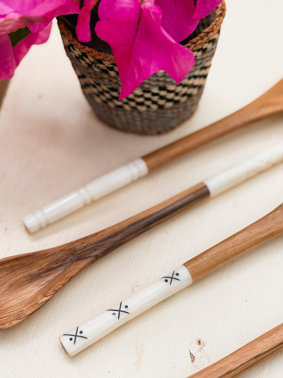 Olive Wood Sugar & Honey Spoons with white decorative handles, including one with black X patterns, are displayed on a light surface beside a woven basket filled with bright pink flowers.