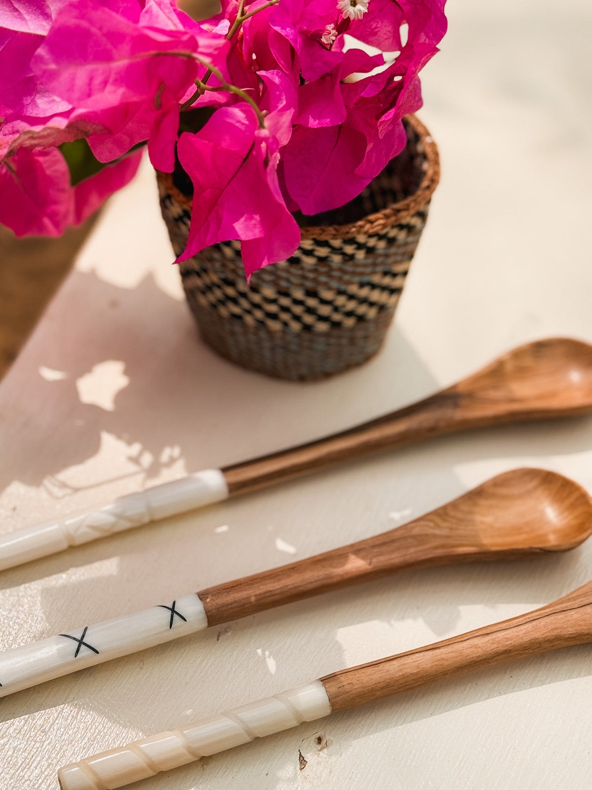 Three Olive Wood Sugar & Honey Spoons, featuring white and black handle details, are displayed on a light background beside a woven basket with pink flowers—unique Kenyan artisan utensils for honey or sugar.