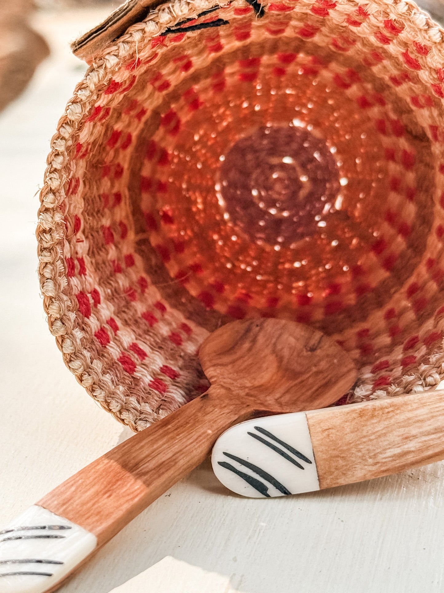 A woven basket with red and natural patterns lies on its side, with two Mini Olive Wood Spice Spoons—featuring white handles and black line designs—placed in front, all arranged on a light surface.