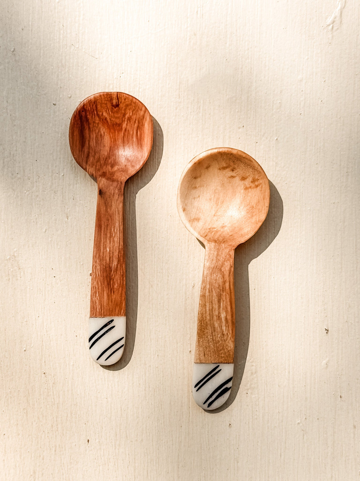 Two Mini Olive Wood Spice Spoons with rounded bowls and white-tipped handles, each accented by black diagonal stripes, are displayed on a light textured surface with gentle shadows.