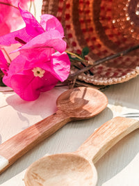 A close-up of Mini Olive Wood Spice Spoons beside vibrant pink bougainvillea and a woven basket. Sunlight casts soft shadows, highlighting the charm of handmade Nairobi Kenya crafts.