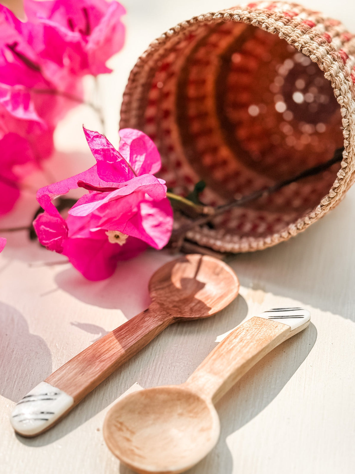A woven basket tipped over with vibrant pink bougainvillea and two Mini Olive Wood Spice Spoons, each with white and black painted tips, all set on a light surface in bright sunlight—showcasing Nairobi Kenya craftsmanship.