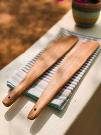 Two handmade olive wood serving tongs rest on a folded striped cloth atop a white surface. In the background, a partially visible colorful woven basket with flowers showcases Nairobi, Kenya craft traditions.