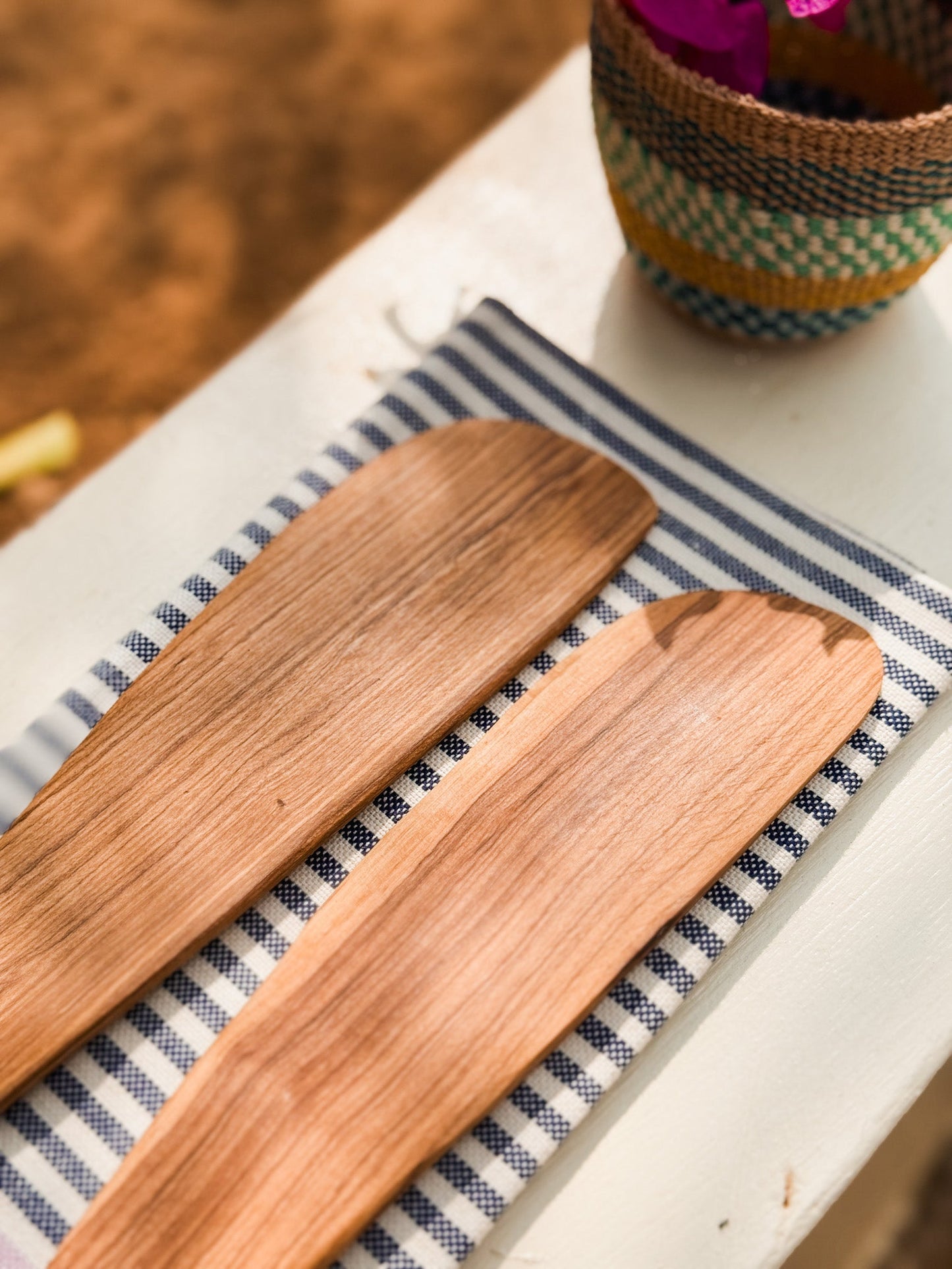 Two Olive Wood Serving Tongs rest side by side on a blue and white striped cloth, with part of a woven basket filled with pink flowers visible in the upper right corner—a charming touch of Nairobi Kenya crafts.