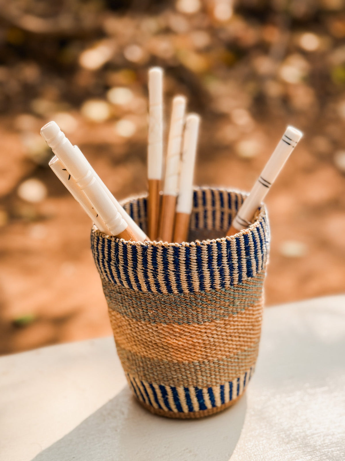 A woven, striped cup holds several Olive Wood Sugar & Honey Spoons, crafted with Kenyan artisan style, displayed on a light surface against a softly blurred outdoor background.