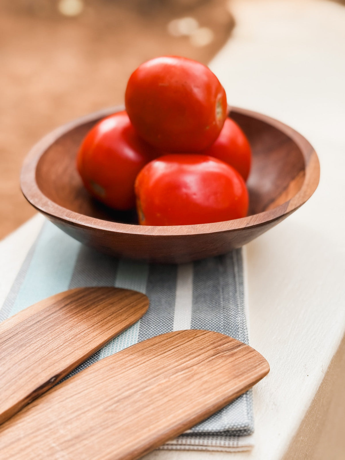 A wooden bowl with four red tomatoes sits on a striped cloth beside handmade kitchen utensils and the Olive Wood Serving Tong Set, all arranged on a white surface with a blurred outdoor background.