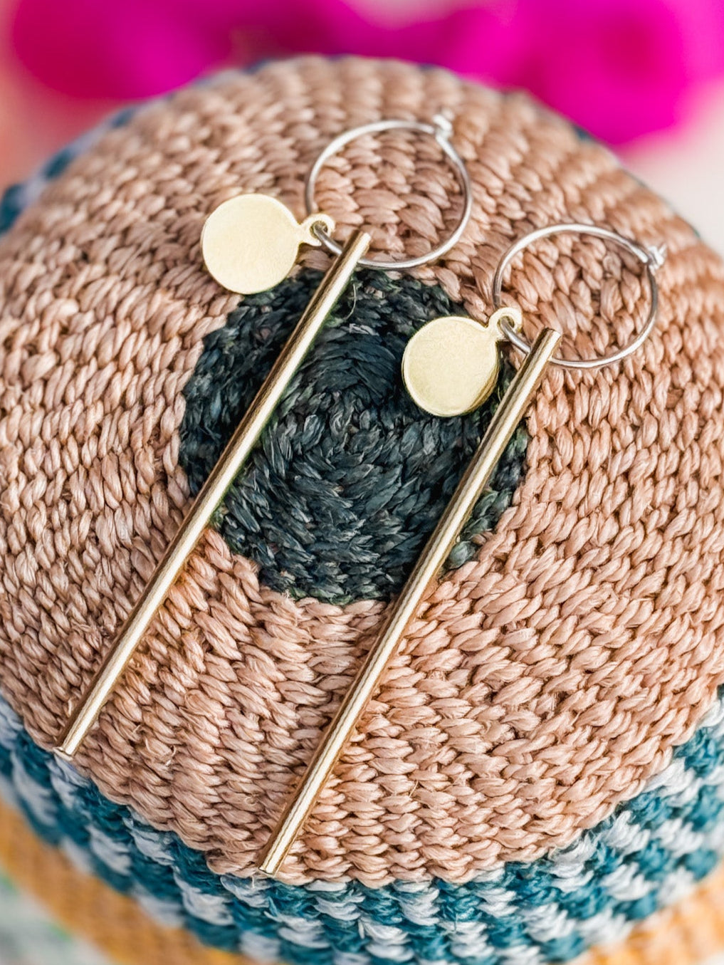 The Rising Sun Earrings feature thin gold vertical bars and small round discs, elegantly displayed on a textured crocheted background with concentric multicolored rings—a stylish handmade jewelry piece.