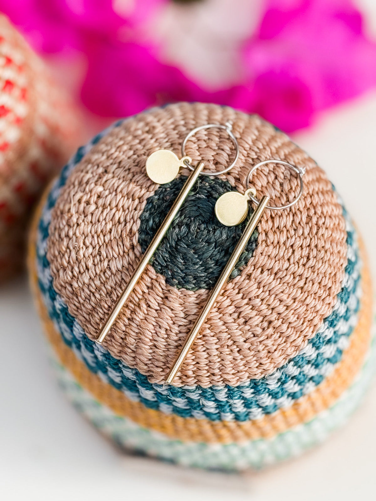 The Rising Sun Earrings—minimalist gold jewelry with circular studs and slender bars—are displayed on a round, woven multicolored basket, with pink flowers softly blurred in the background.