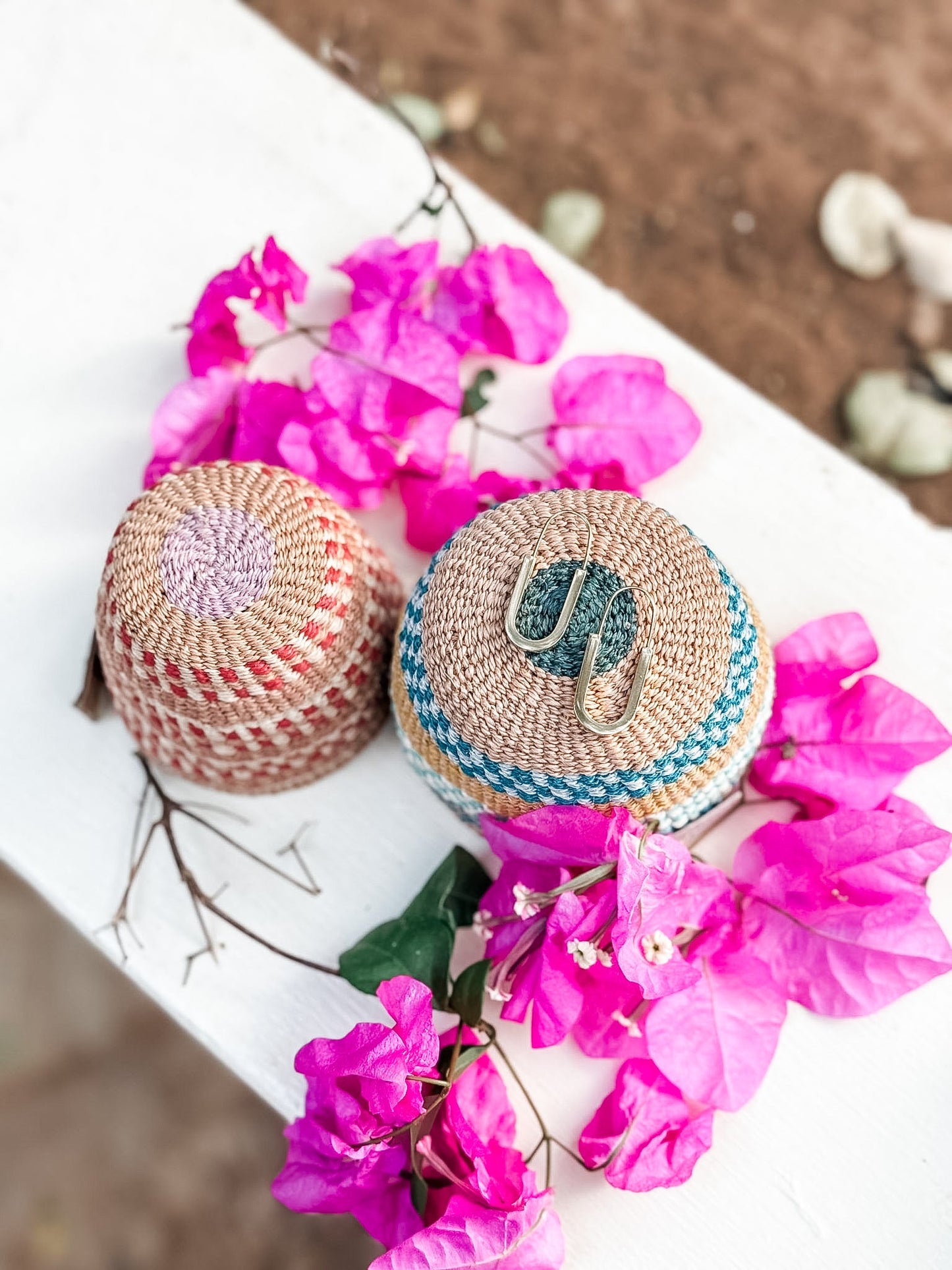 Two woven baskets—one pink, one blue—hold a pair of Drop Out Earrings, surrounded by vibrant pink bougainvillea on a white outdoor surface. Perfect for showcasing unique South African jewelry and mixed-metal charm earrings.