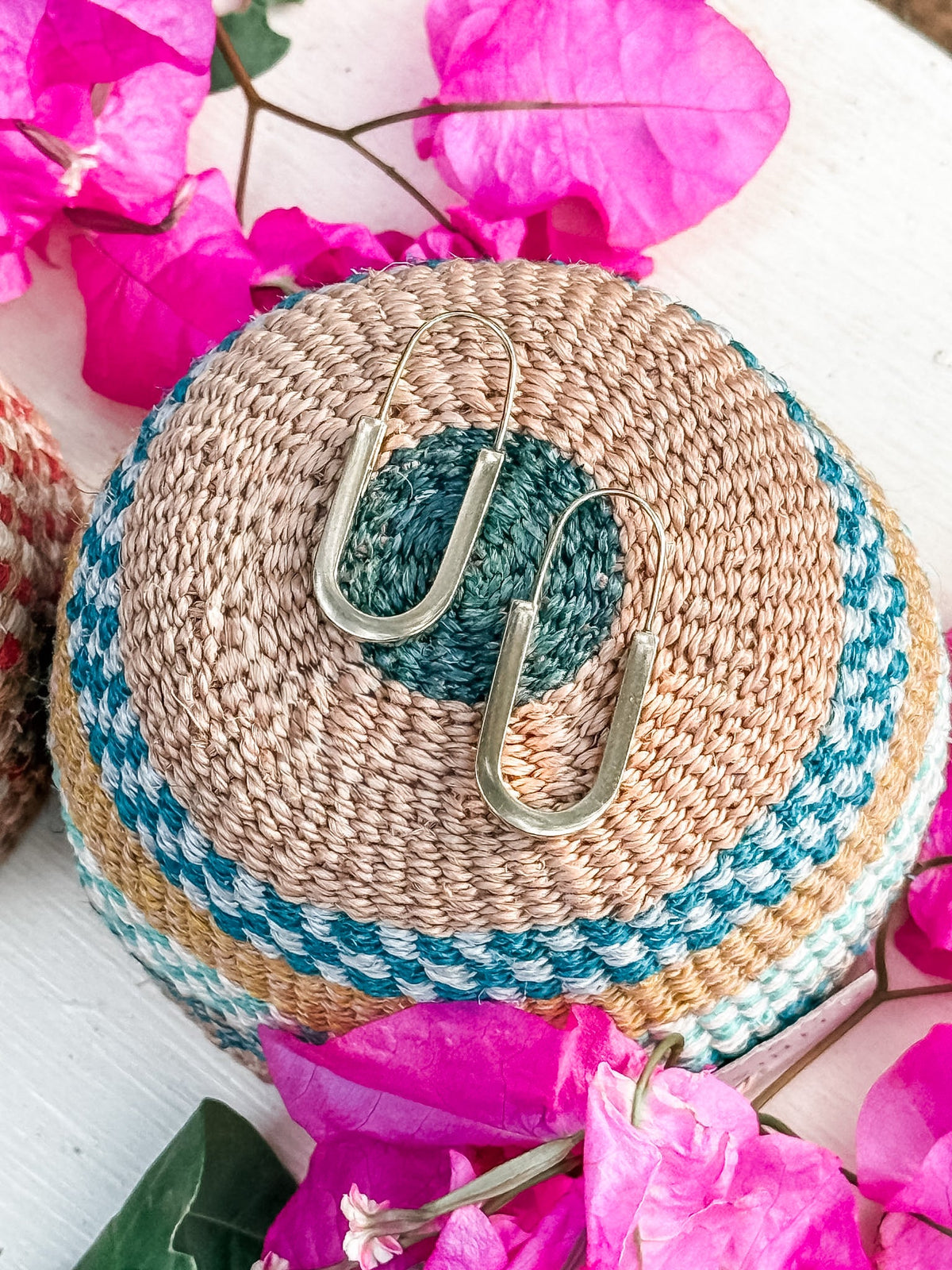 The Drop Out Earrings, a handmade gold U-shaped pair, are displayed on a woven multicolored basket amid vibrant pink bougainvillea flowers.