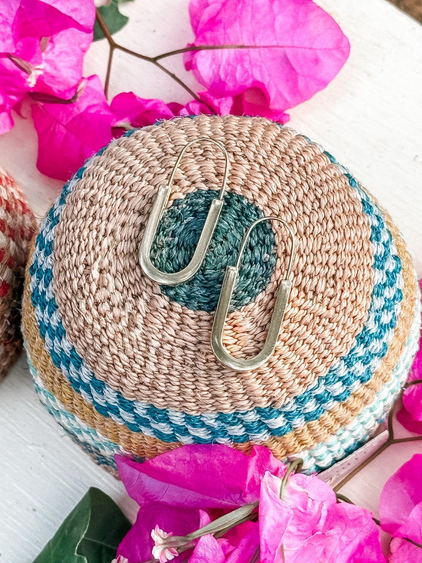 The Drop Out Earrings, a handmade gold U-shaped pair, are displayed on a woven multicolored basket amid vibrant pink bougainvillea flowers.