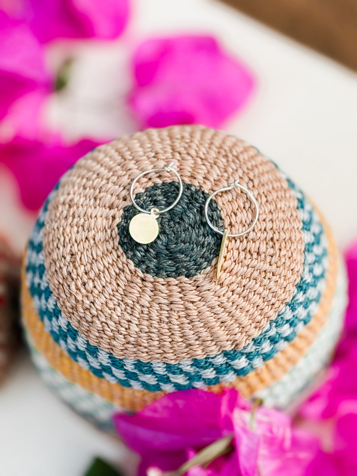 The Akabeko Earrings, minimalist gold handmade jewelry, rest on a woven multicolored basket lid amid bright pink bougainvillea—a stunning example of Cape Town style.