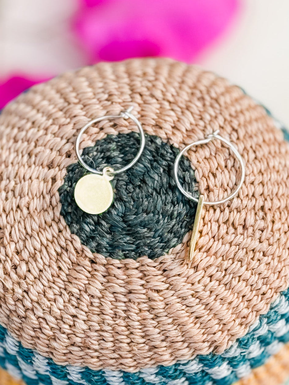The Akabeko Earrings—handmade from brass and sterling silver—rest on a woven, multicolored basket. One earring has a round gold charm, the other a slim bar, with soft pink hues in the background highlighting this Cape Town jewelry.