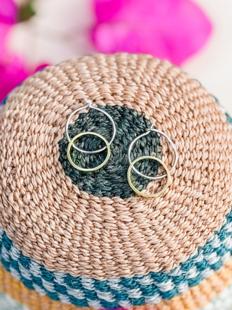 The Hello Halo Earrings, a small mixed-metal pair, rest on a round woven basket lid with tan, black, and blue concentric patterns. Blurred pink flowers in the background highlight the artisanal charm of Cape Town jewelry.