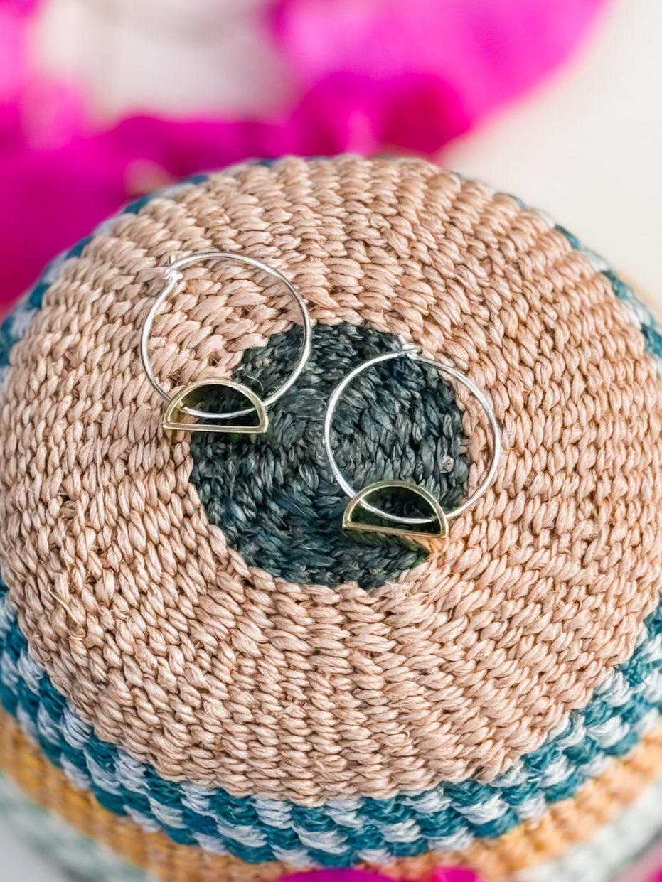 The Setting Sun Earrings—silver hoops with gold semicircle accents, handmade in Cape Town—rest on a round woven basket with tan, blue, and green patterns. Bright pink fabric is softly blurred in the background.