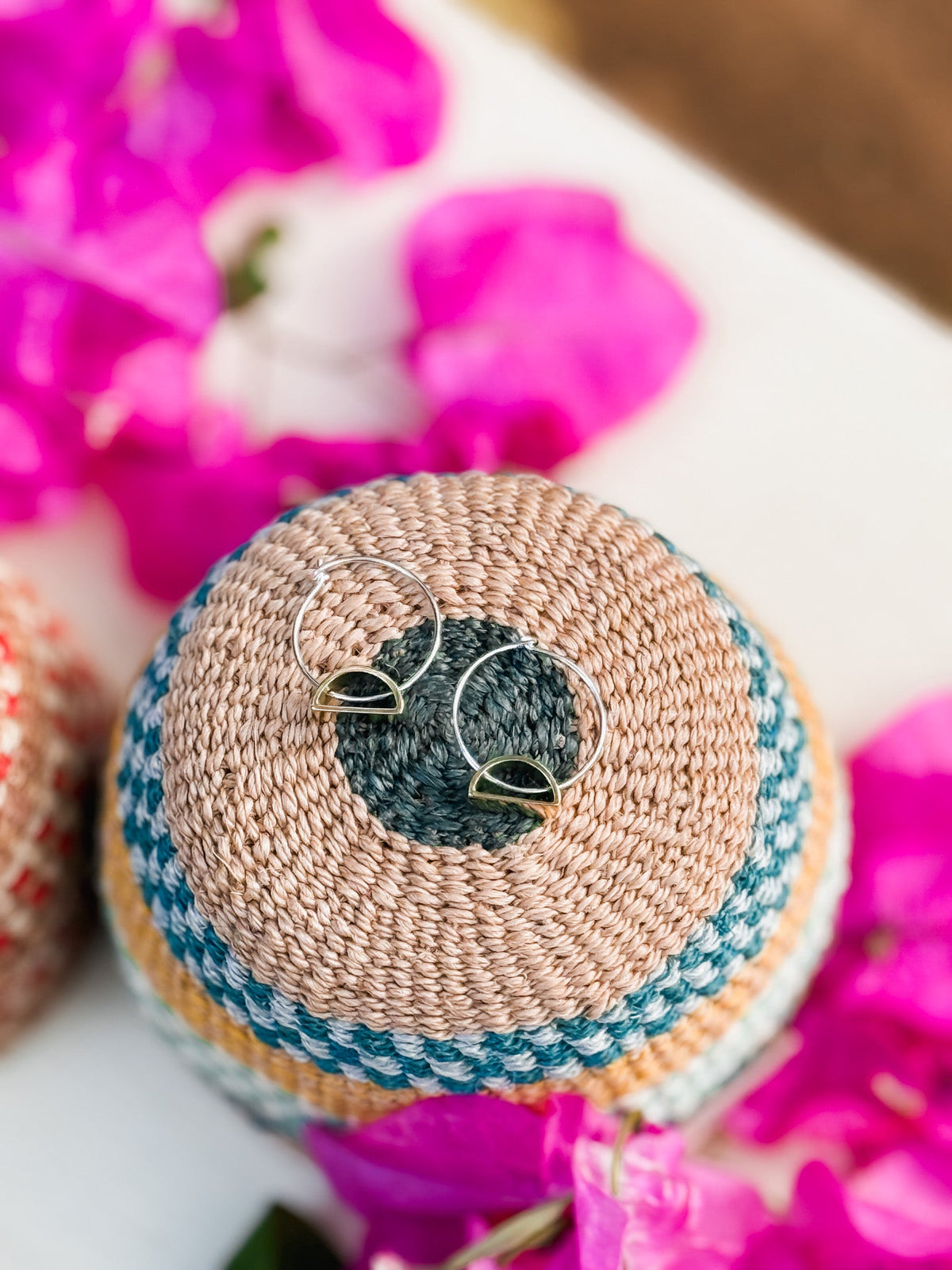 The Setting Sun Earrings, handmade silver hoops with black and gold accents, rest on a woven multicolored basket surrounded by bright pink bougainvillea—a striking piece of Cape Town jewelry.