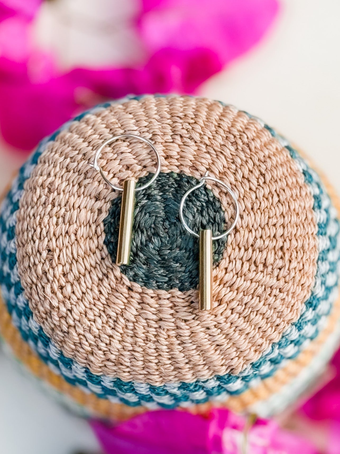 Golden Rays Earrings, handmade gold cylinder hoops, are displayed on a woven round basket with tan, green, and yellow sections and surrounded by softly blurred pink flowers.