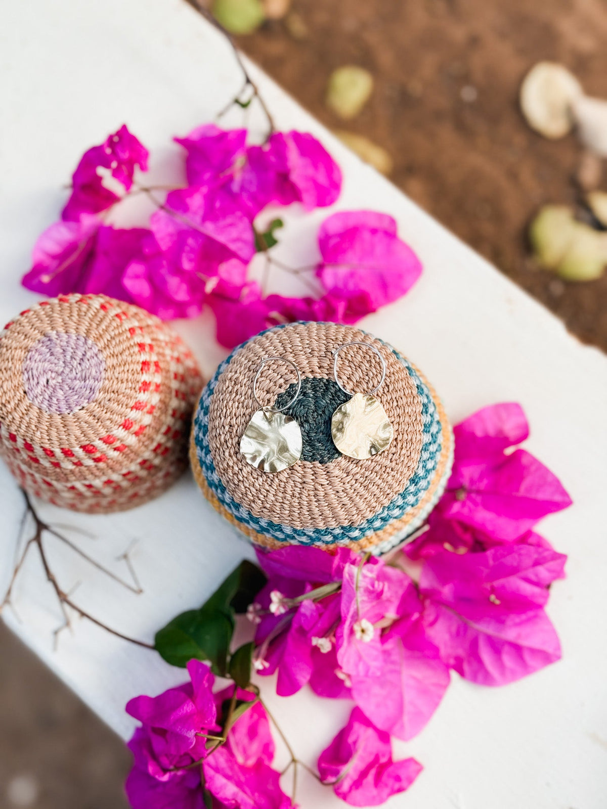 Two woven baskets—one striped, one with blue accents—sit on a white surface with pink bougainvillea. The blue-accented basket holds a pair of Autumn Leaves Earrings, featuring mixed metals and handmade craftsmanship.