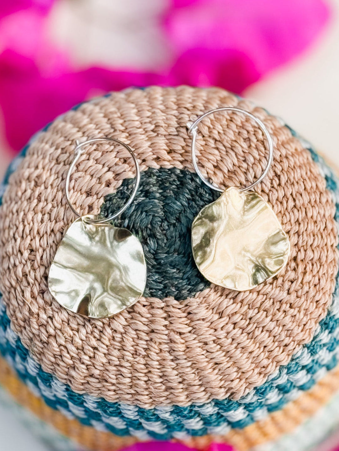 The Autumn Leaves Earrings are gold, textured, disc-shaped handmade jewelry with hoop fastenings, displayed on a woven multicolored basket. Blurred pink flowers appear in the background.