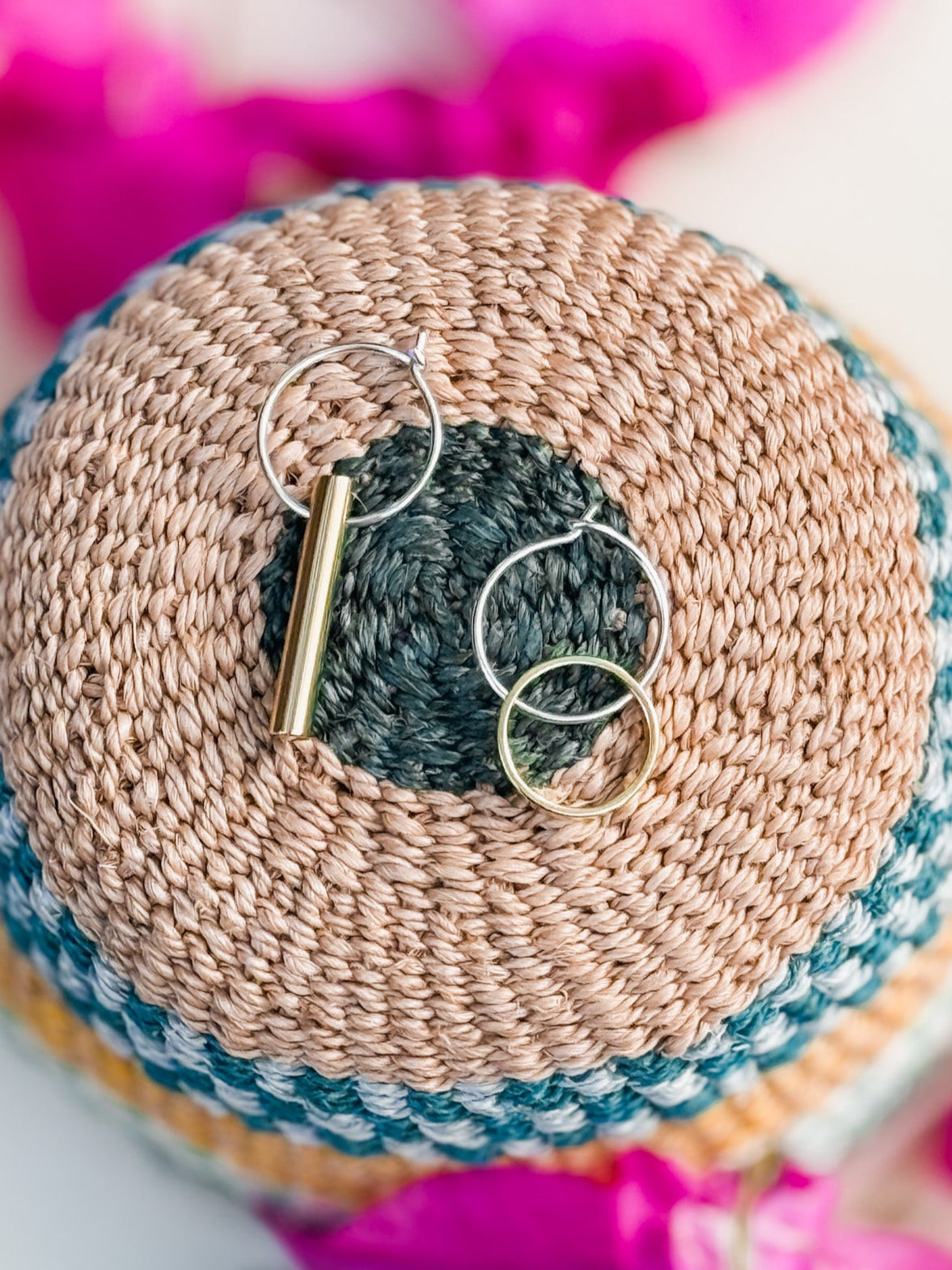 The Hello Halo & Ray Earrings—one a cylindrical bar, the other a simple hoop—are displayed on a colorful woven round mat in Cape Town, accented with pink flower petals.