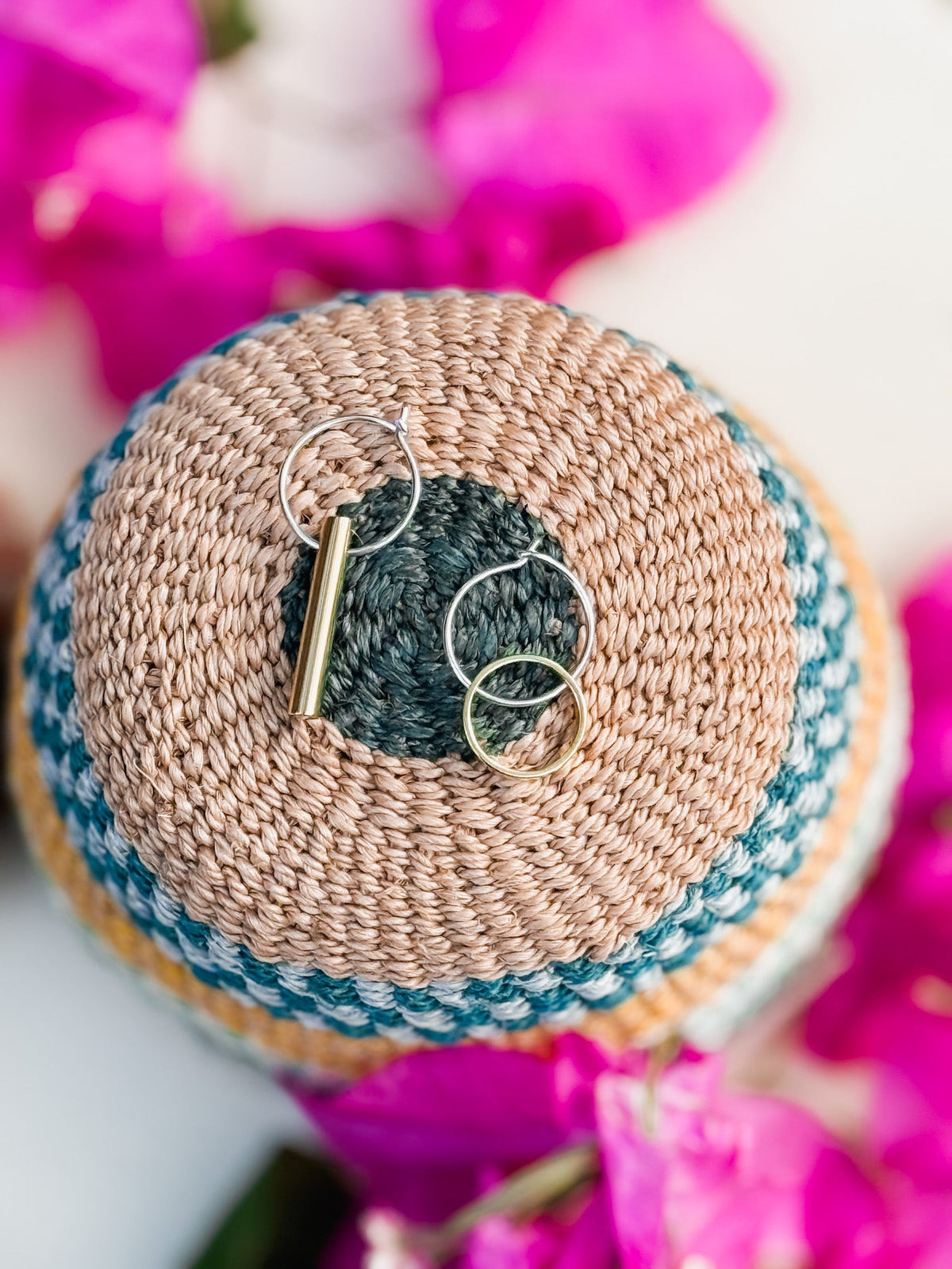 The Hello Halo & Ray Earrings, a pair of handmade mixed-metal earrings—one with a gold bar charm—are displayed on a round woven basket with beige, green, and blue stripes, surrounded by bright pink flowers.