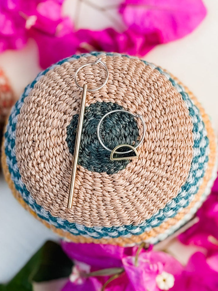 A woven, round basket lid in teal and natural tones displays the Falling Light & Setting Sun Earrings, surrounded by vibrant pink bougainvillea petals.