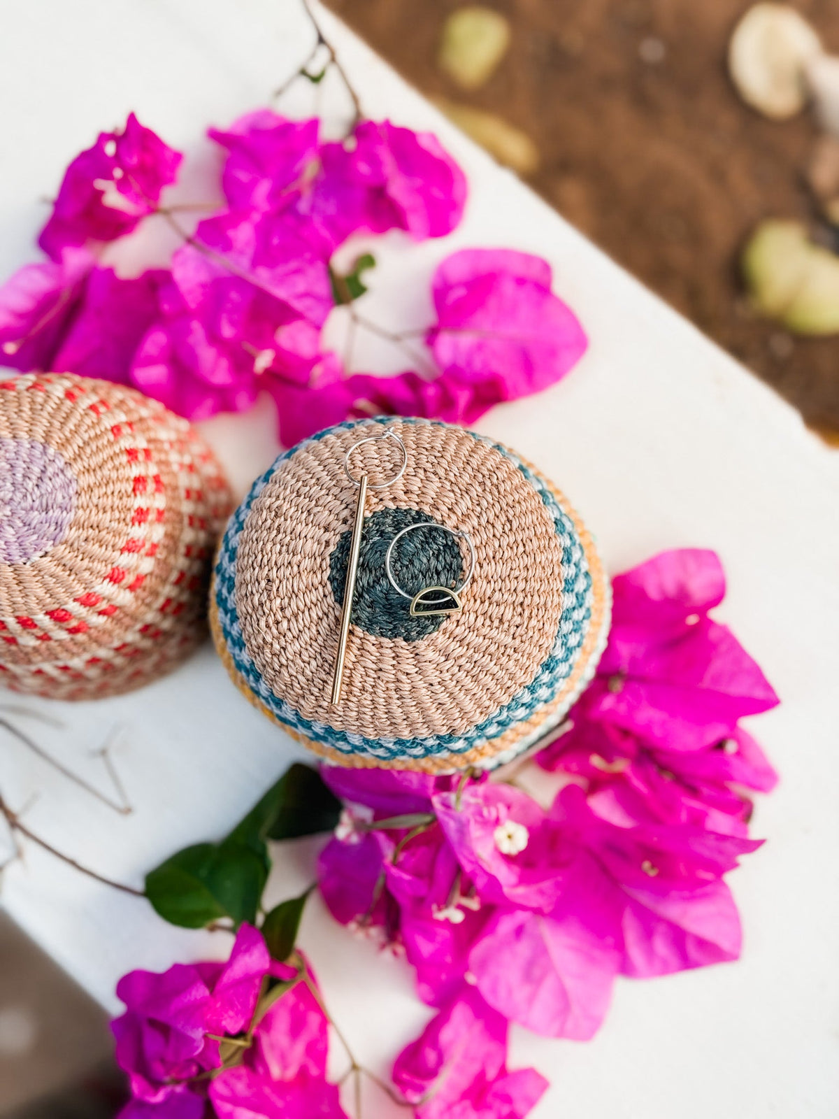 The Falling Light & Setting Sun Earrings rest on a woven basket lid, surrounded by vibrant pink bougainvillea on a white surface with a soft natural background—a shining example of Cape Town jewelry craftsmanship.