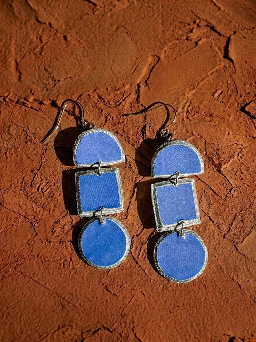 A pair of Lindiwe Paper Pulp Earrings by Quazi Designs rests on a textured brown surface, featuring blue semicircle, square, and circle shapes with silver edges—handcrafted from recycled newspaper.