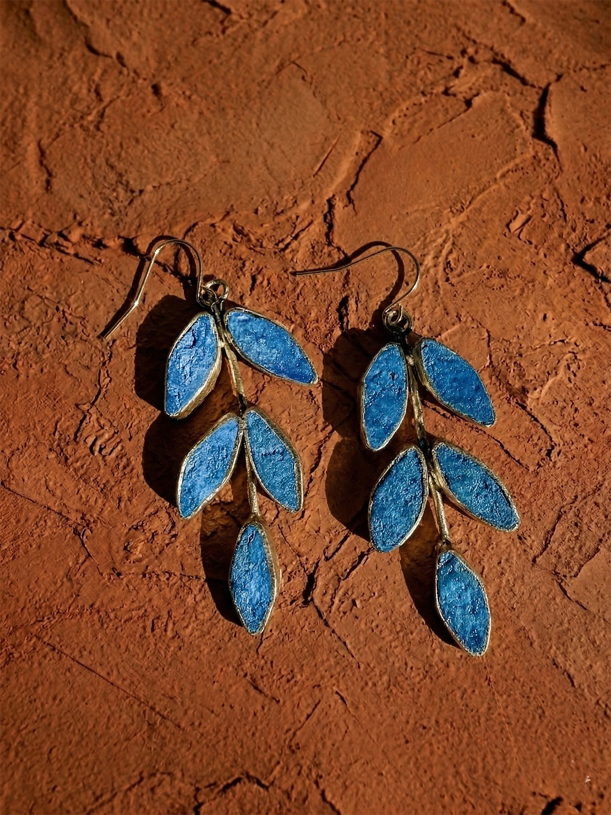 The Posy Paper Pulp Earrings are handcrafted and branch-shaped with blue leaf-like segments, shown on a textured brown surface.