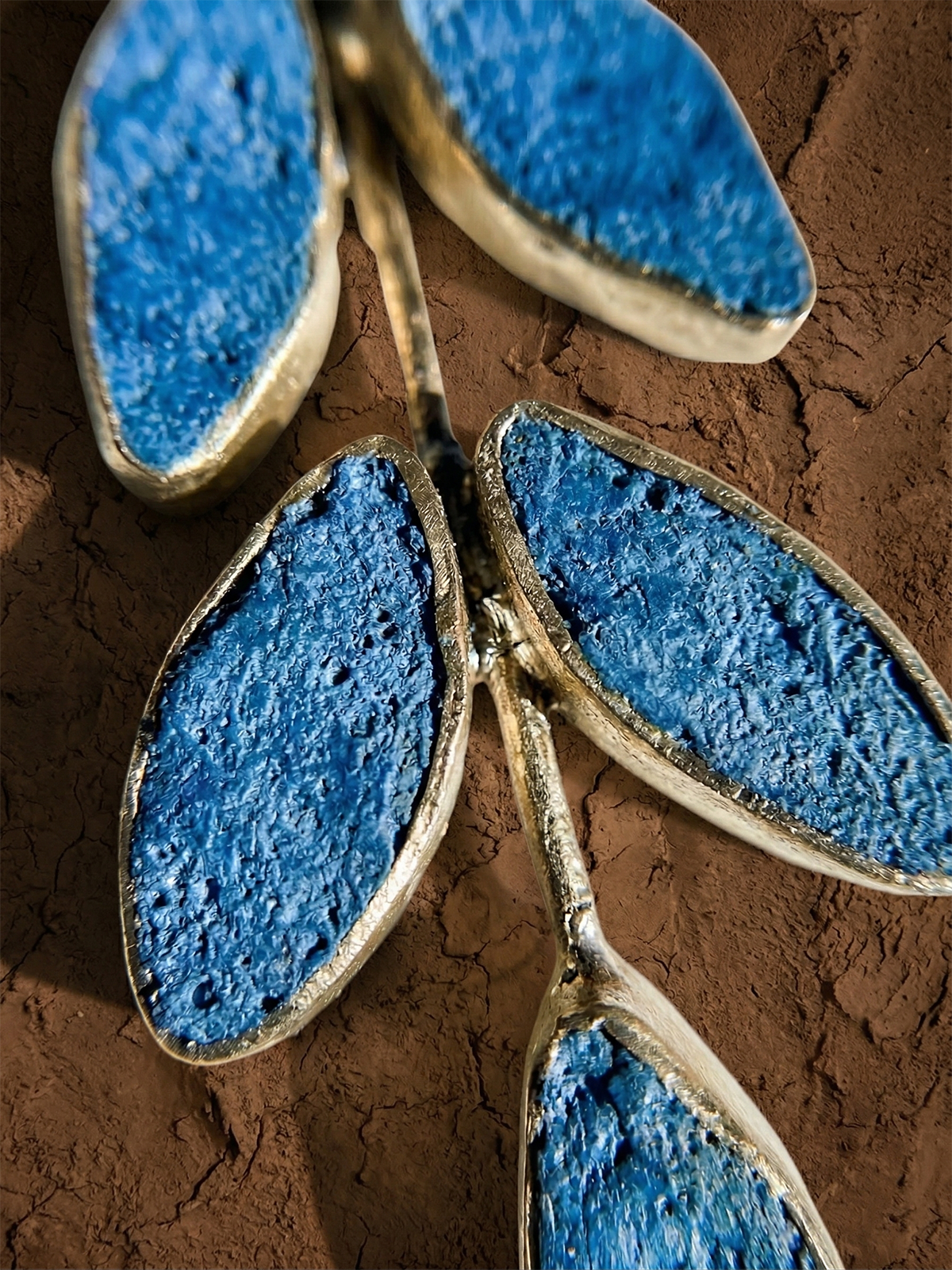 Close-up of Posy Paper Pulp Earrings: decorative metal branches with blue, textured, oval leaves edged in metallic silver on a brown, cracked surface. The rough, stone-like leaves evoke handcrafted artistry.