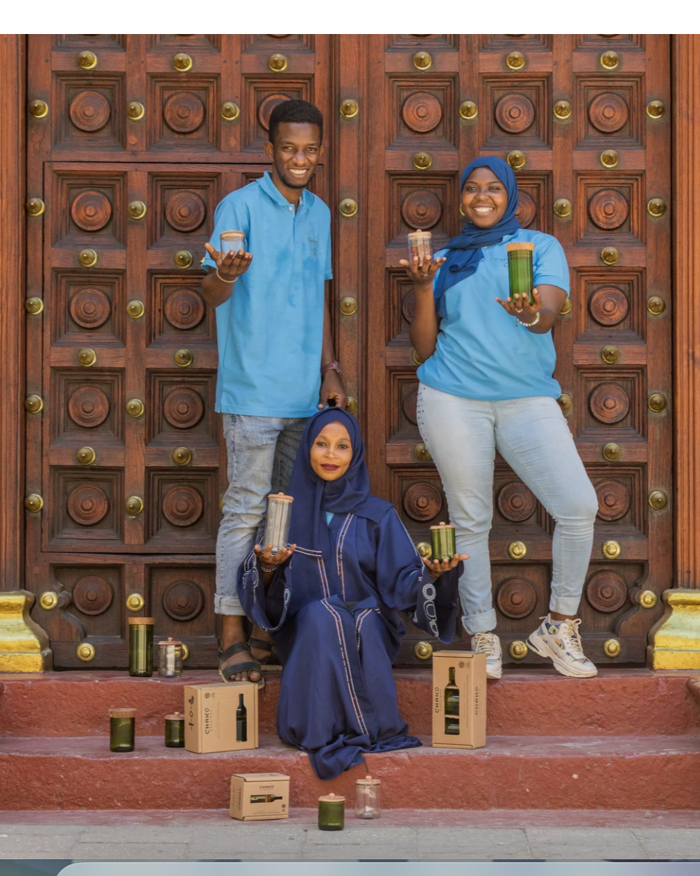 Three people smile in front of an ornate wooden door, holding candles from the Hiliki Upcycled Glass Jars | Large Set. They are surrounded by jars and boxes with beautifully carved wooden lids arranged on the steps around them.