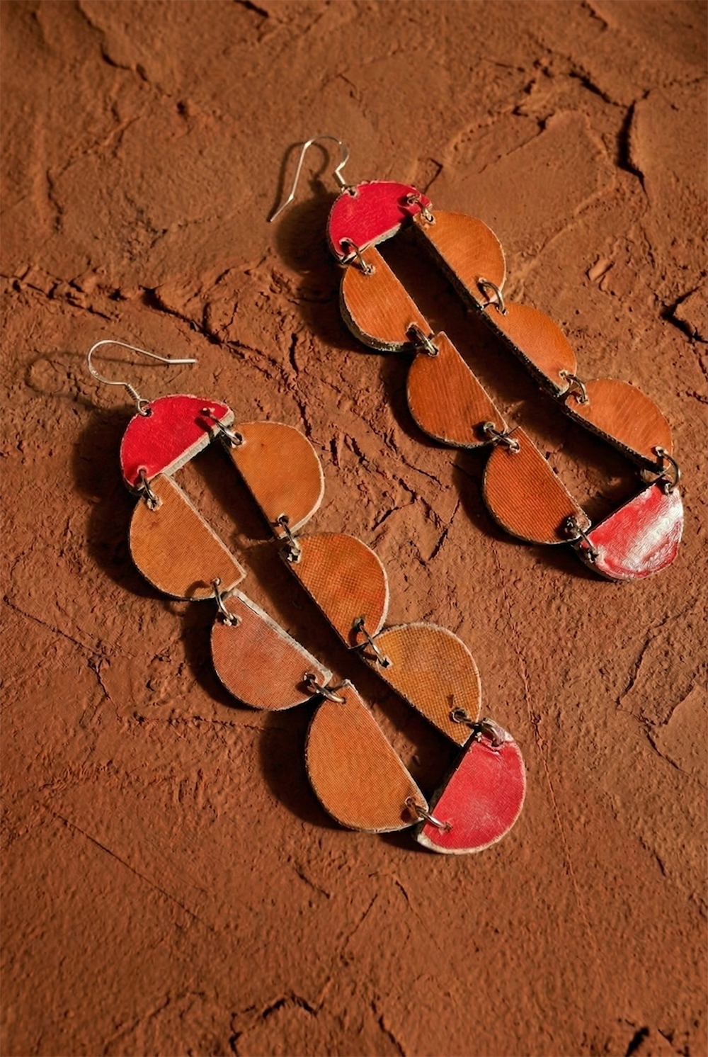 The Sisanda Paper Pulp Earrings by Quazi Design are handcrafted long dangles made from recycled newspaper in linked brown and red semi-circles, shown on a textured reddish-brown surface.