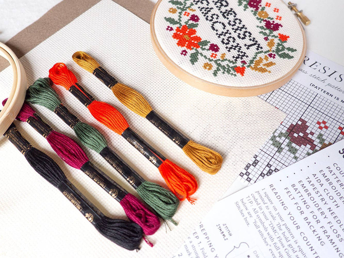 The Resist Fascism Cross Stitch Kit includes colorful embroidery threads, a needle, hoop, pattern, and instructions to stitch "RESIST FASCISM" above a floral border—perfect for beginners.