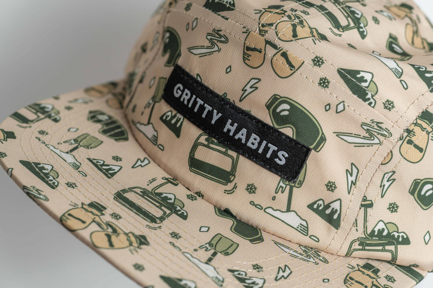 Close-up of the water-resistant First Chair 5-Panel Hat in beige, featuring green outdoor-themed illustrations and a black patch with white GRITTY HABITS text. Made from performance polyester.
