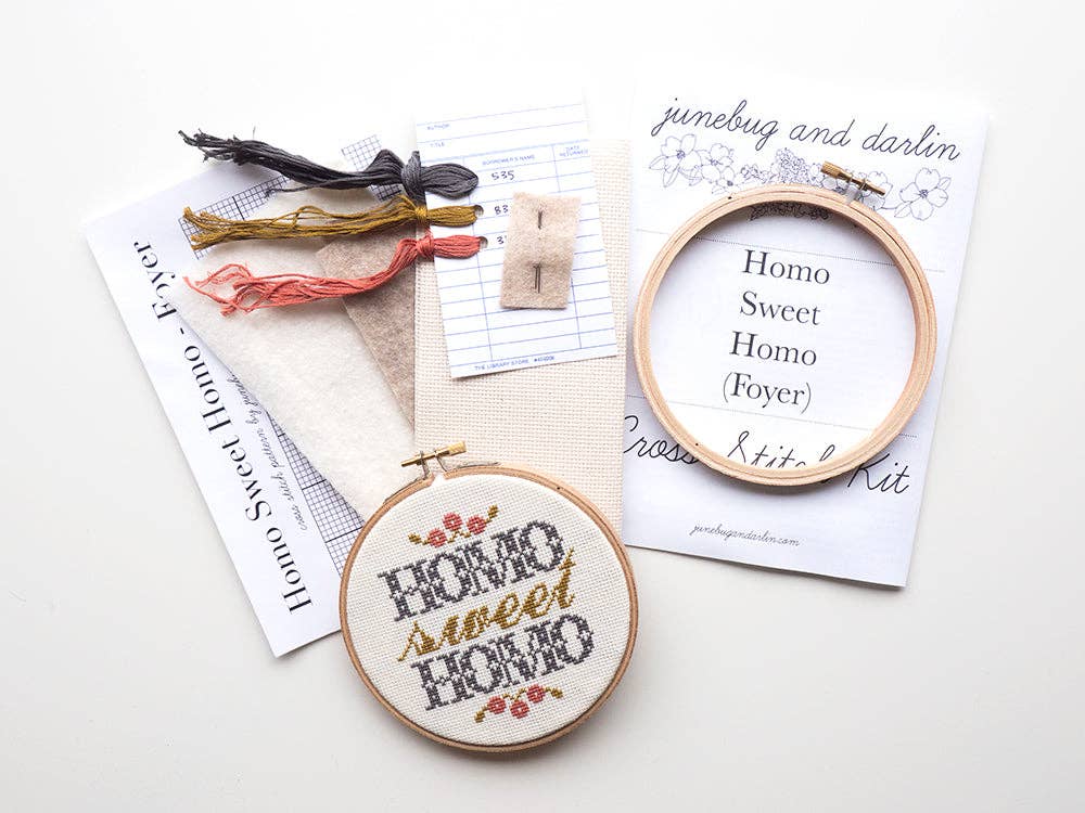 The Homo Sweet Homo (Foyer) Cross Stitch Kit includes threads, fabric, needle, instructions, and a hoop. It creates a floral "Homo sweet homo" design—perfect as a Pride Month gift or for beginners learning cross stitch.