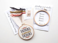The Homo Sweet Homo (Foyer) Cross Stitch Kit includes threads, fabric, needle, instructions, and a hoop. It creates a floral "Homo sweet homo" design—perfect as a Pride Month gift or for beginners learning cross stitch.