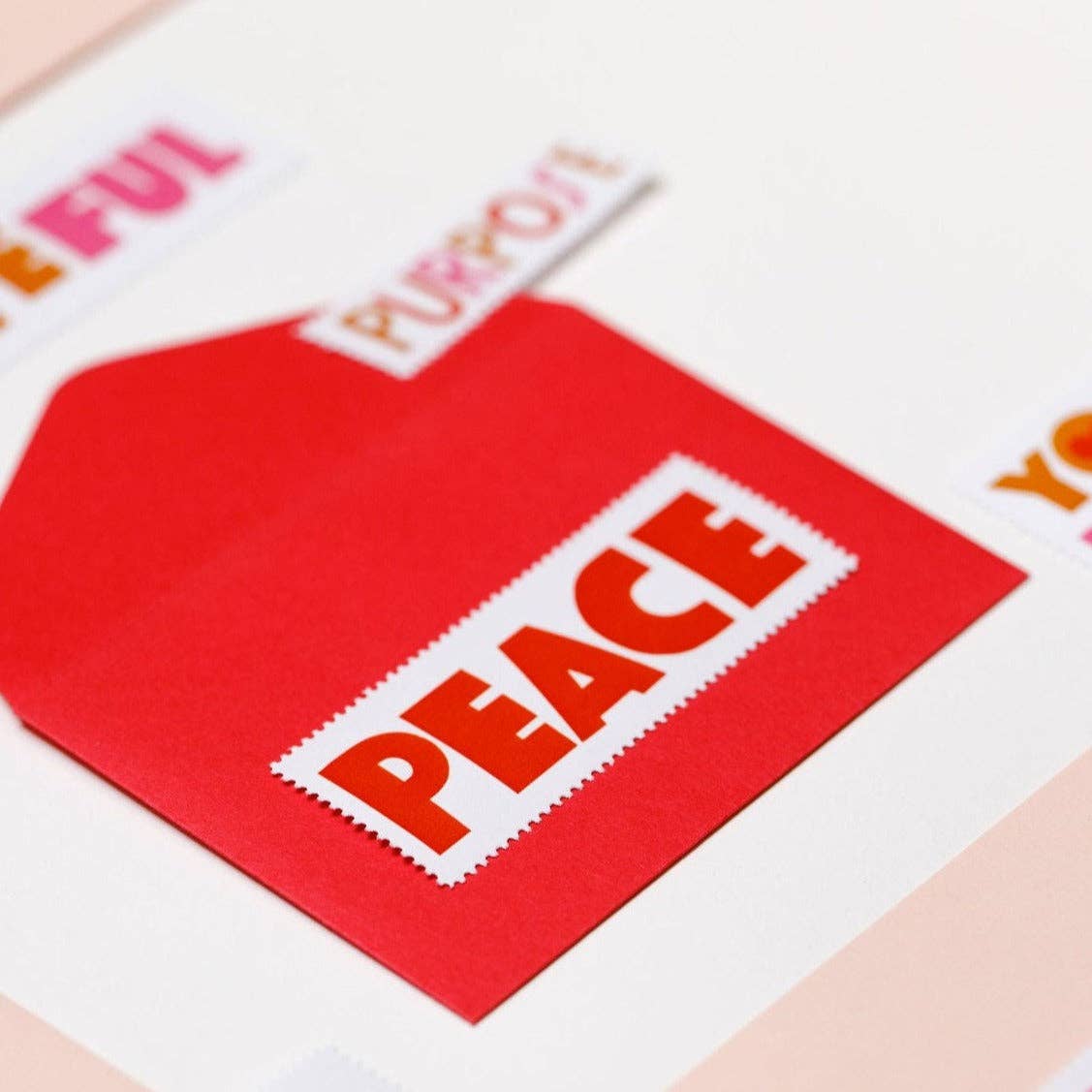 A red envelope rests on a white surface with a white label reading PEACE in bold red letters. Cutout words like PURPOSEFUL and PURPOSE, from Poster Stamp Series 5 | Words to Live By, are also visible.