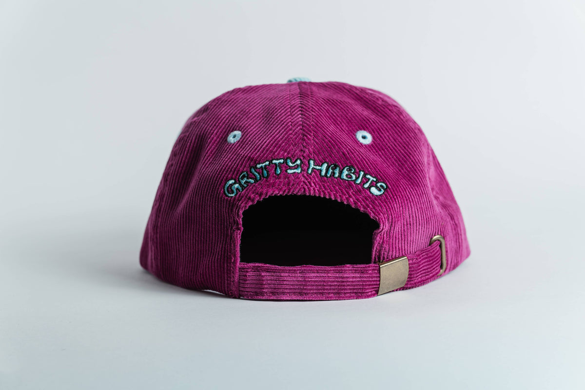 The Shred Til She's Dead Corduroy 5-Panel Hat in magenta is shown from the back, displaying GRITTY HABITS embroidery above the adjustable strap, plus a moisture-wicking sweatband for comfort.