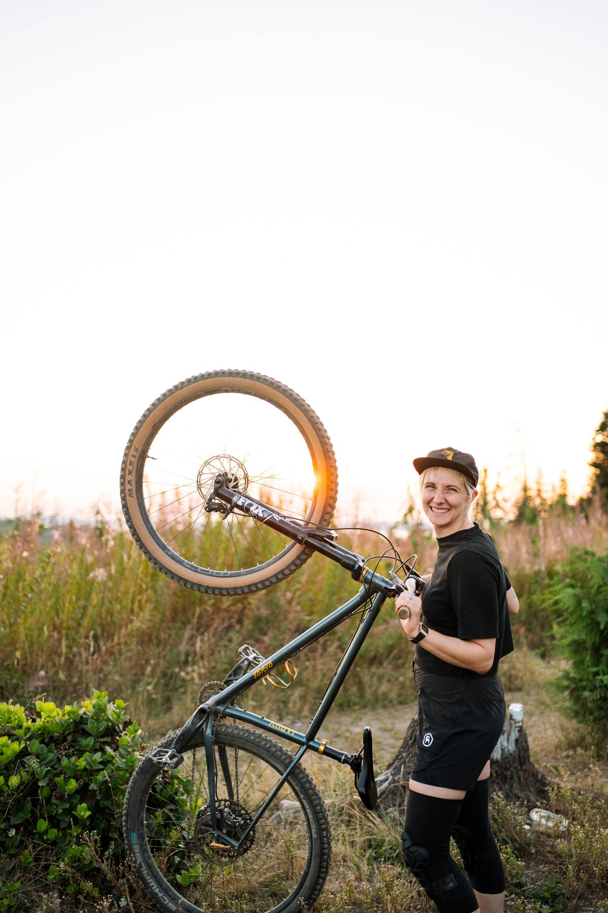 Wearing a Fearless 5-Panel Hat from Gritty Habits, a person in outdoor gear smiles while lifting a mountain bike upright on its rear wheel in a grassy field at sunset.