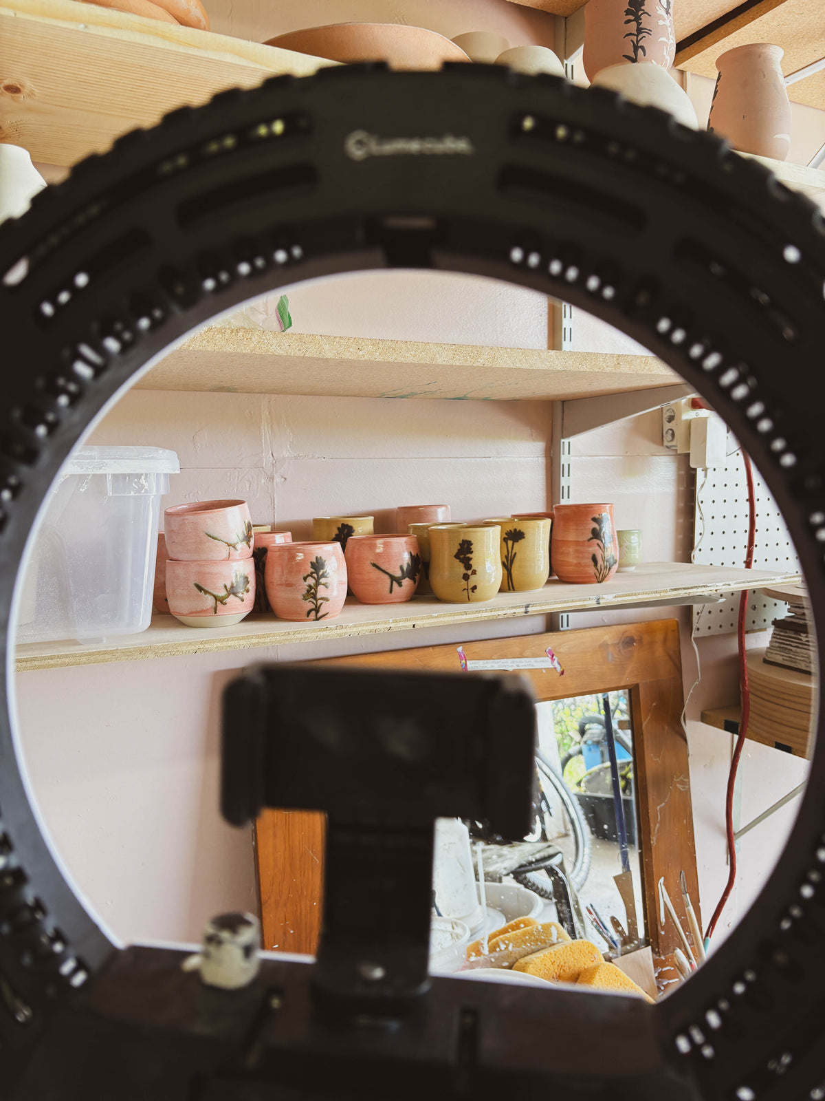 A ring light with a phone holder frames a view of the Birch Arnica #1 Small Tea Cup, a handcrafted ceramic piece with painted designs, displayed on a wooden shelf in a pottery studio as outdoor greenery reflects in the window.