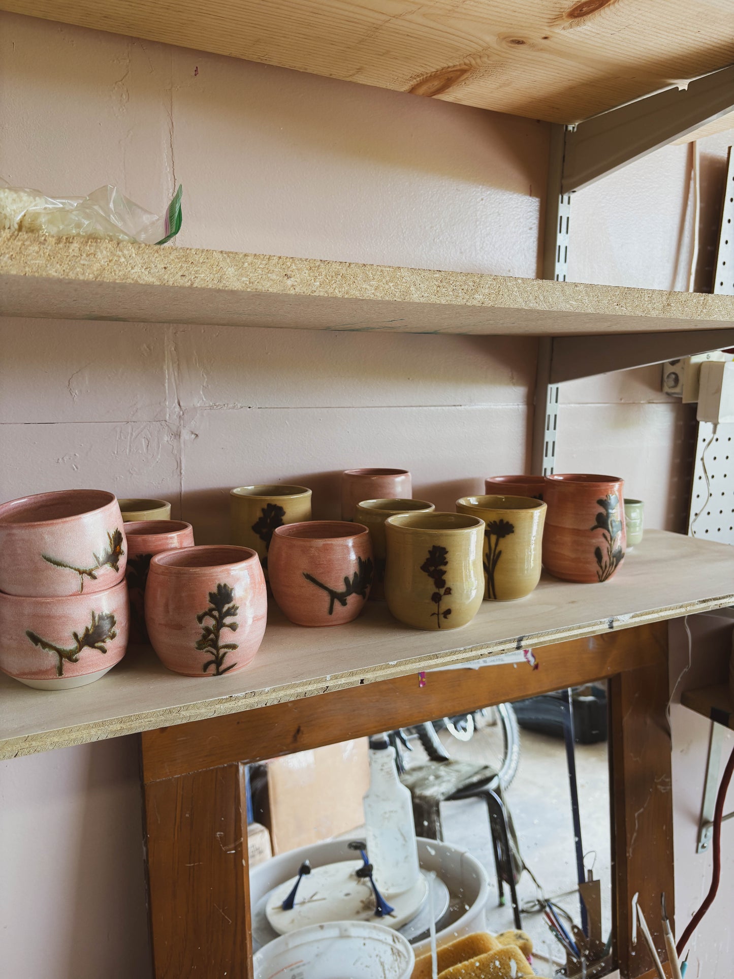 The Birch Globemallow #2 Medium Tea Cup, a handcrafted ceramic cup in pink and yellow with black floral designs, is displayed on a wooden shelf in a pottery studio with shelving, walls, and pottery tools in the background.