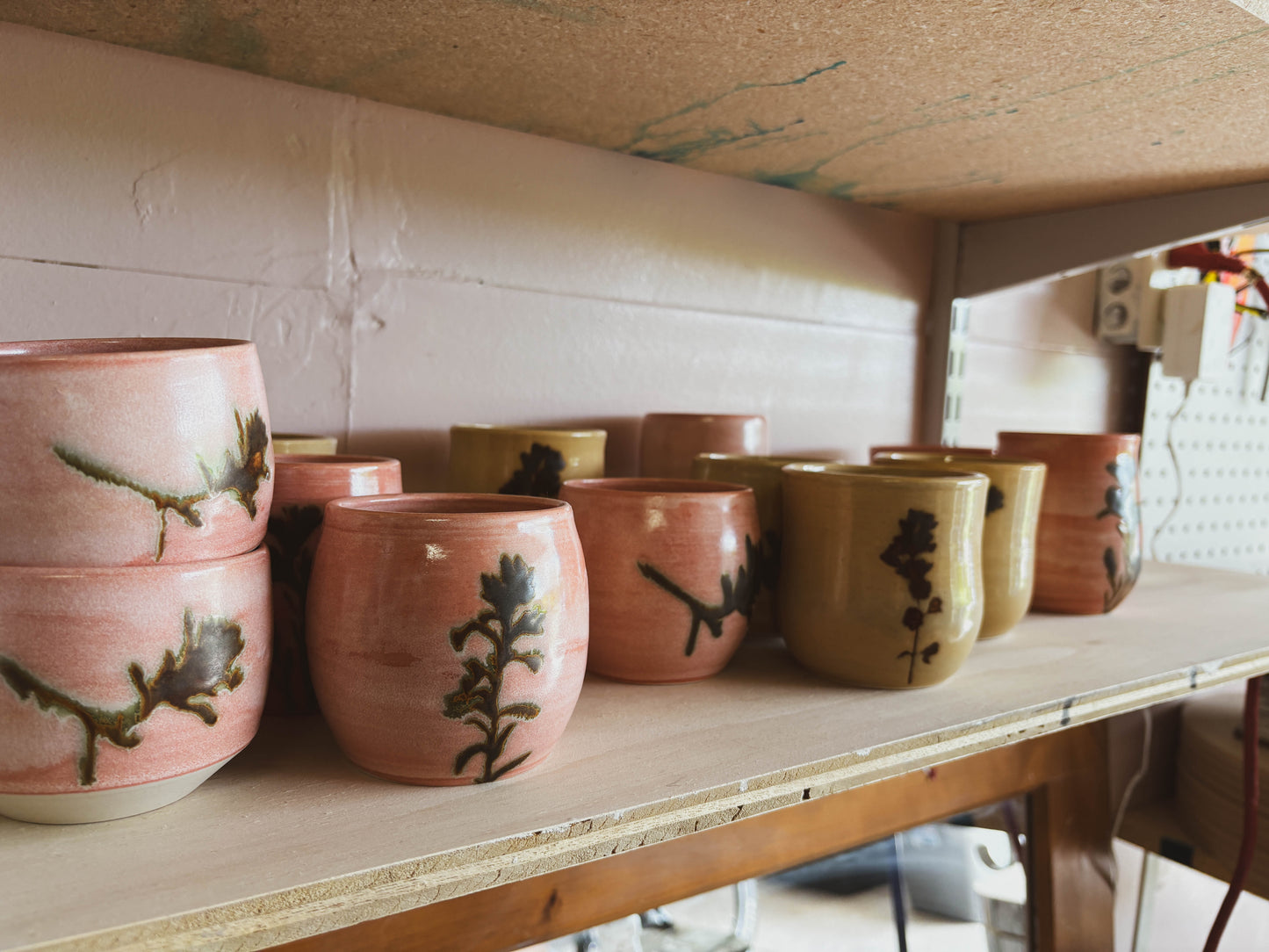 The Rose Quartz Paintbrush #4 Small Tea Cup, handcrafted in a women-owned studio with pink and tan glazes and leaf designs, is neatly displayed on a wooden shelf—and it's dishwasher safe for easy cleaning.