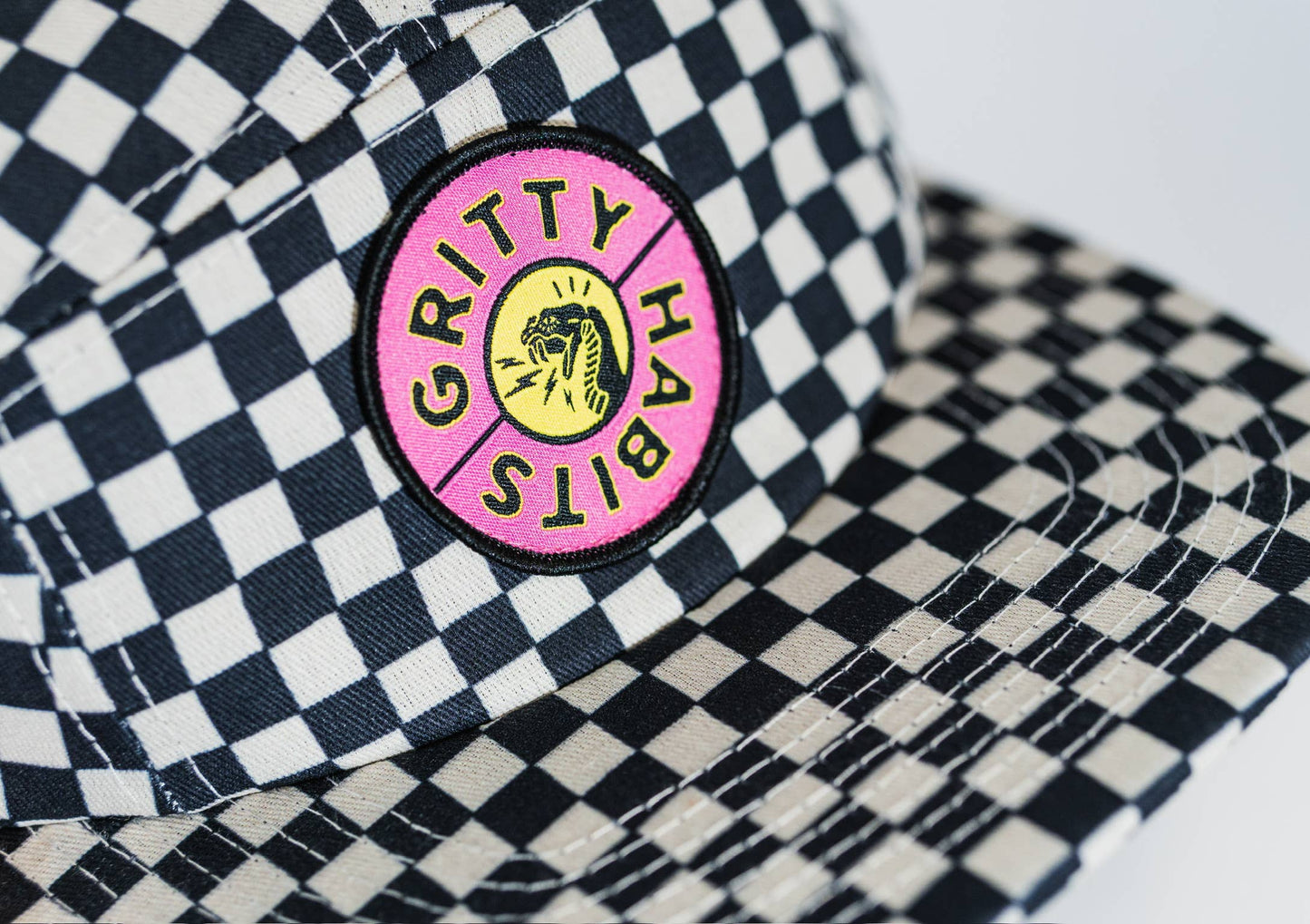 A close-up of the Steezy Checkered 5-Panel Hat, a black-and-white checkered accessory from a women-owned small business, featuring a GRITTY HABITS patch with a roaring tiger in pink, yellow, and black.