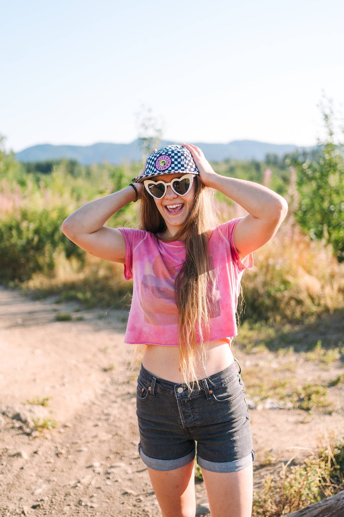 A smiling woman in heart-shaped sunglasses, a Steezy Checkered 5-Panel Hat, pink crop top, and black shorts stands outdoors on a sunny day with trees and mountains in the background.