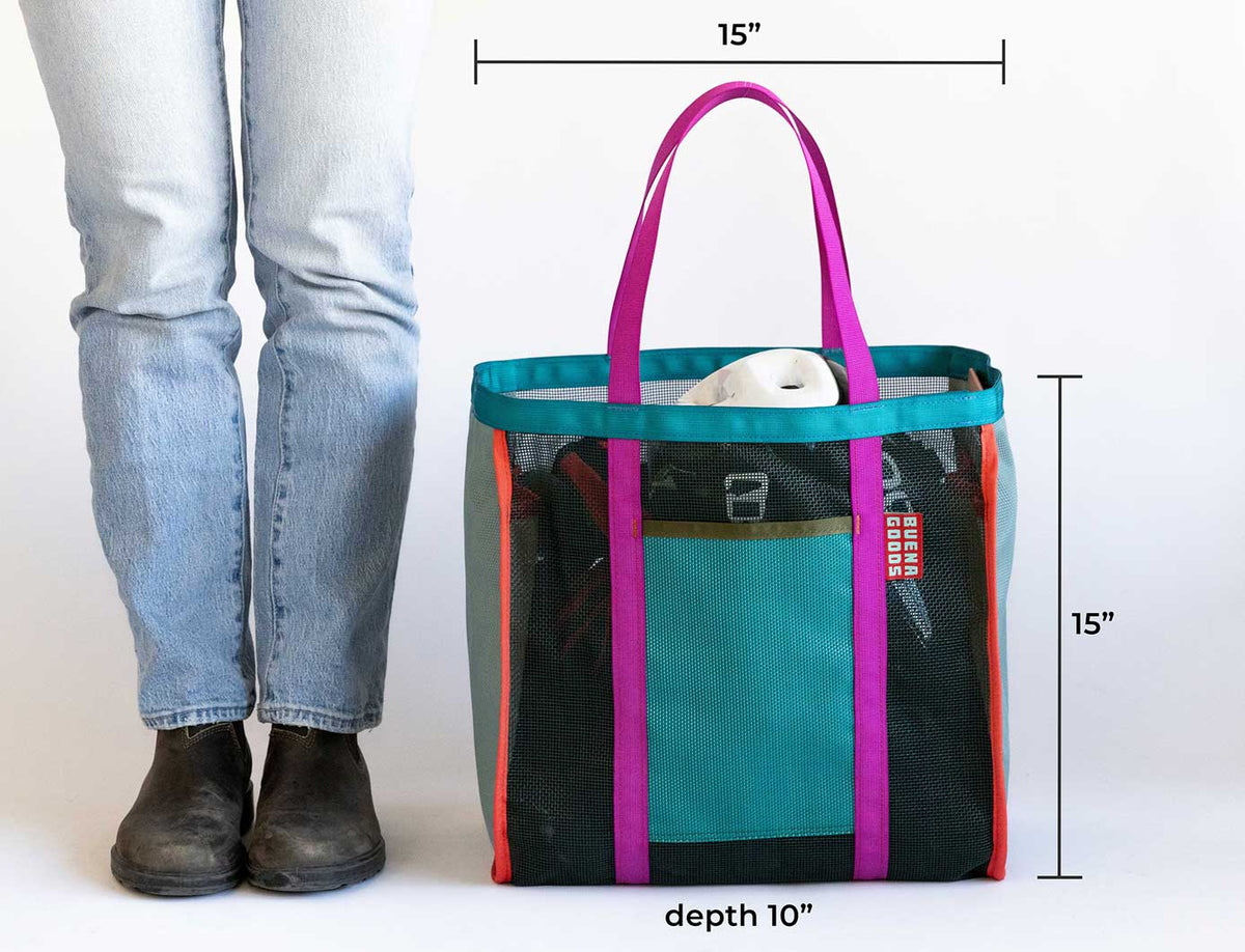 A person in jeans and black boots stands next to The Mother | Yacht Rock, a women-owned large beach tote with purple handles, measuring 15" x 15" x 10", durable mesh design, and roomy enough to carry various items.